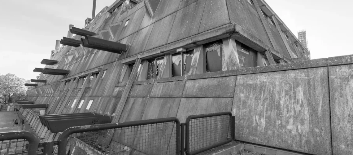 Gebäudeecke des Mäusebunkers in Berlin-Lichterfelde aus der Untersicht: nach außen geneigte Sichtbetonwände mit dreieckigen Fensterelementen, seitlich austretende Luftansaugrohre, Absperrgitter im Vordergrund