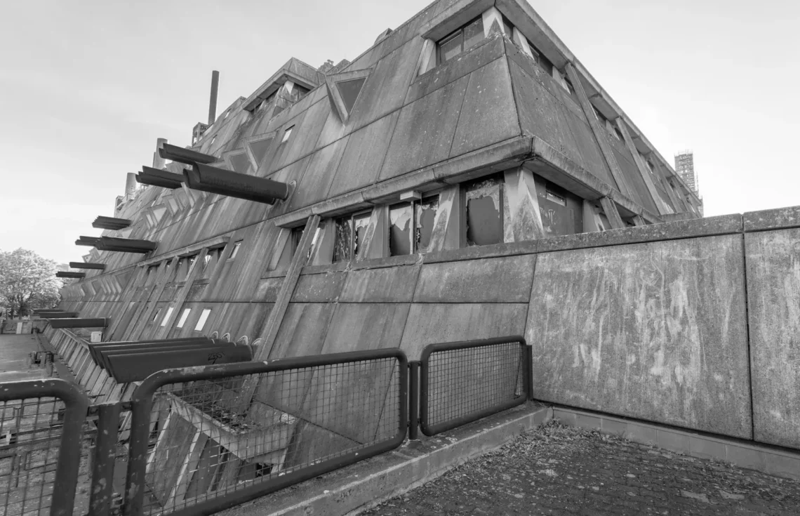 Gebäudeecke des Mäusebunkers in Berlin-Lichterfelde aus der Untersicht: nach außen geneigte Sichtbetonwände mit dreieckigen Fensterelementen, seitlich austretende Luftansaugrohre, Absperrgitter im Vordergrund
