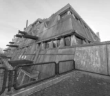 Gebäudeecke des Mäusebunkers in Berlin-Lichterfelde aus der Untersicht: nach außen geneigte Sichtbetonwände mit dreieckigen Fensterelementen, seitlich austretende Luftansaugrohre, Absperrgitter im Vordergrund