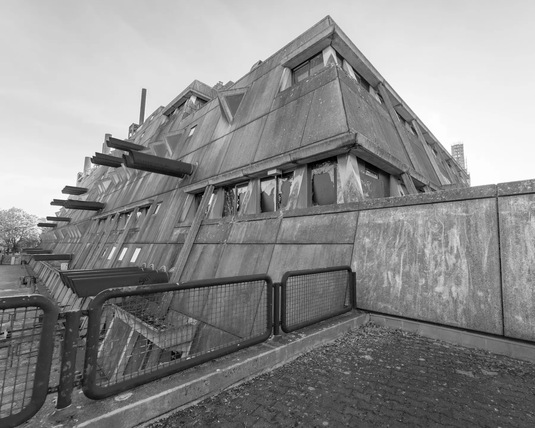 Gebäudeecke des Mäusebunkers in Berlin-Lichterfelde aus der Untersicht: nach außen geneigte Sichtbetonwände mit dreieckigen Fensterelementen, seitlich austretende Luftansaugrohre, Absperrgitter im Vordergrund