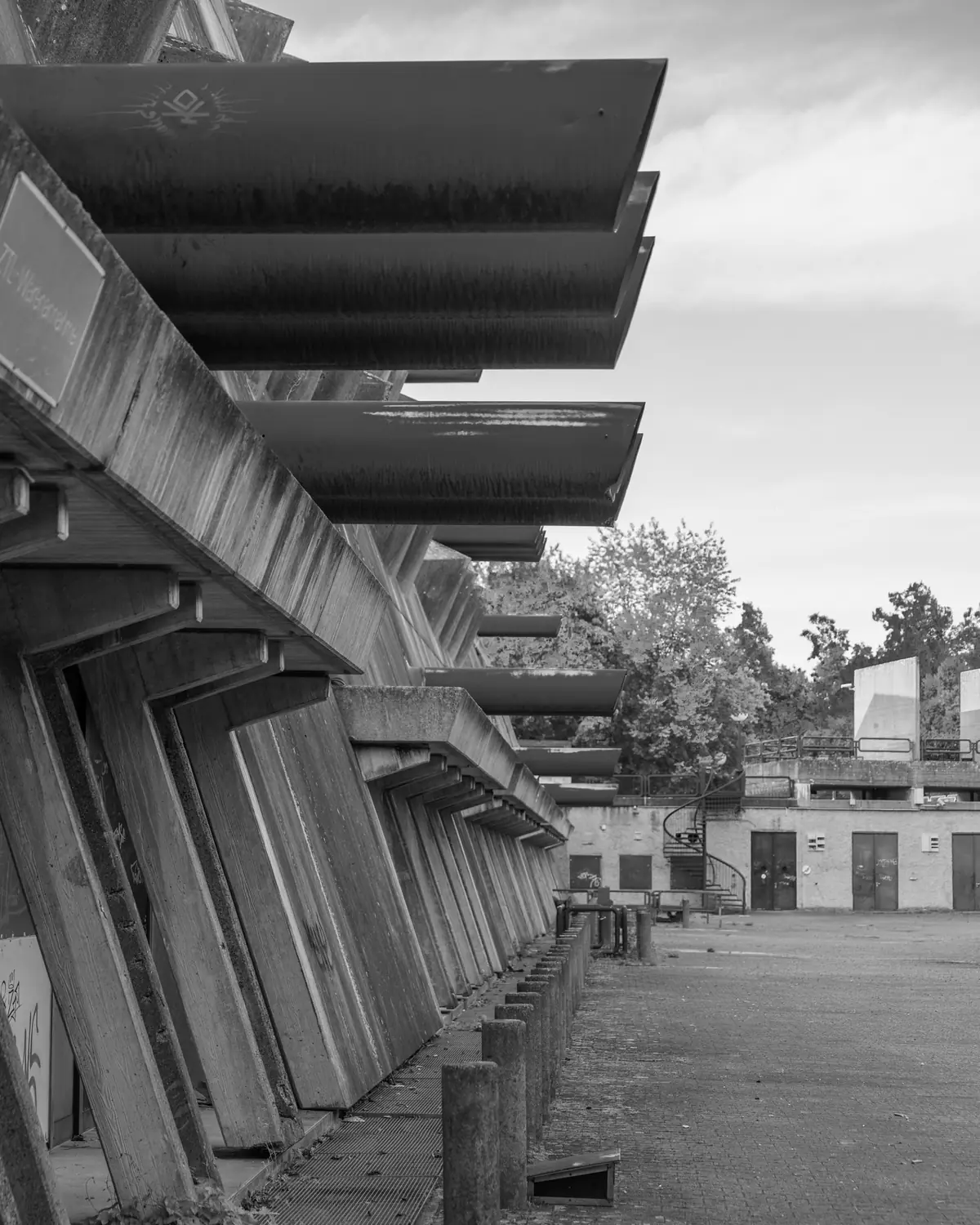 Reihe der Luftansaugrohre an der Längsseite des Mäusebunkers: die Rohre treten weit aus der geneigten Betonwand und laufen in der Flucht auf den leeren Vorplatz zu, im Hintergrund Bäume und flache Nebengebäude