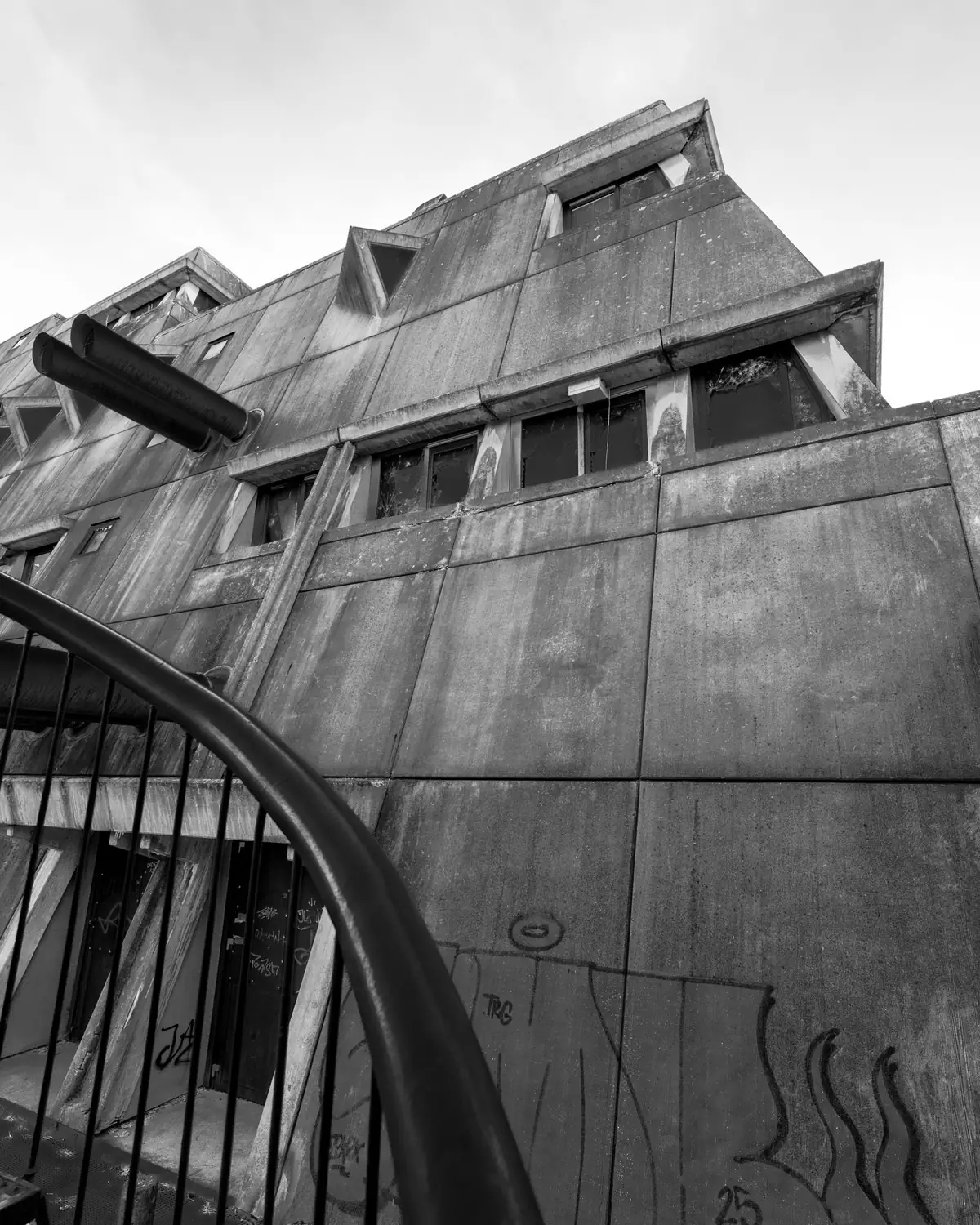 Untersicht auf die Fassade des Mäusebunkers: gestufte Betonwände mit dreieckigen Gauben, aufgebrochene und zerstörte Fensterbänder, Graffiti auf dem Sichtbeton, im Vordergrund ein Geländer