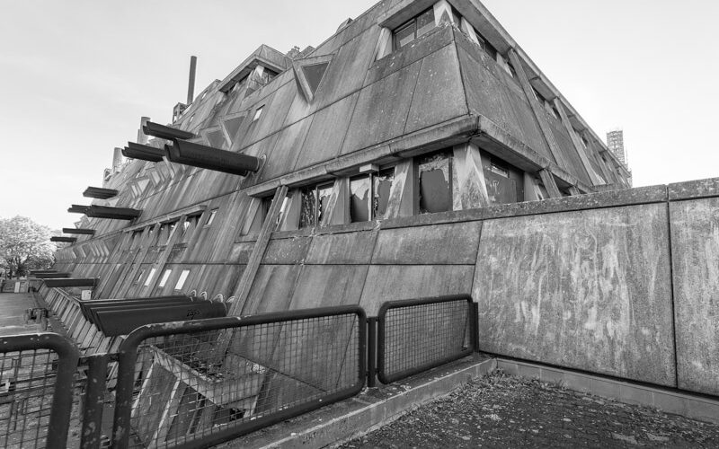 Mäusebunker Berlin-Lichterfelde in scharfer Untersicht: gestufter, nach außen geneigter Sichtbetonbau mit auskragenden Betonstreben und aufgebrochenen Fensterbändern, Absperrung im Vordergrund – Brutalismus, Architekturfotografie in Schwarzweiß