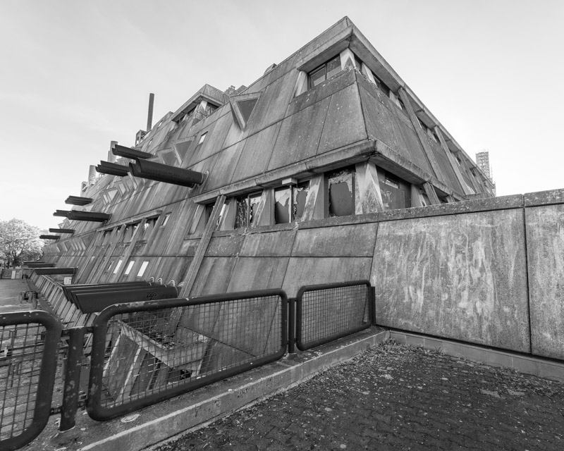 Mäusebunker Berlin-Lichterfelde in scharfer Untersicht: gestufter, nach außen geneigter Sichtbetonbau mit auskragenden Betonstreben und aufgebrochenen Fensterbändern, Absperrung im Vordergrund – Brutalismus, Architekturfotografie in Schwarzweiß