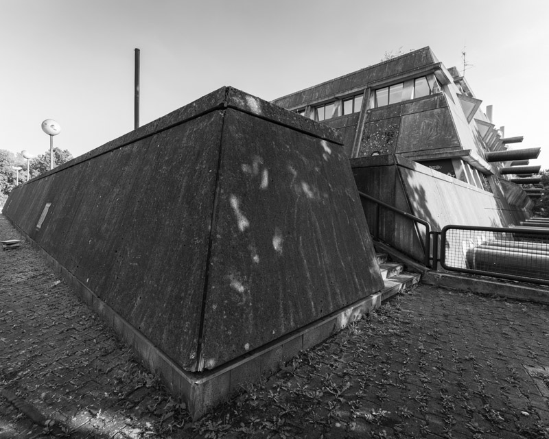 Mäusebunker Berlin: nach außen geneigter Sichtbeton-Baukörper mit trapezförmigem Vorbau, seitlich austretende blaue Luftansaugrohre, Kopfsteinpflaster im Vordergrund – Brutalismus in Schwarzweiß