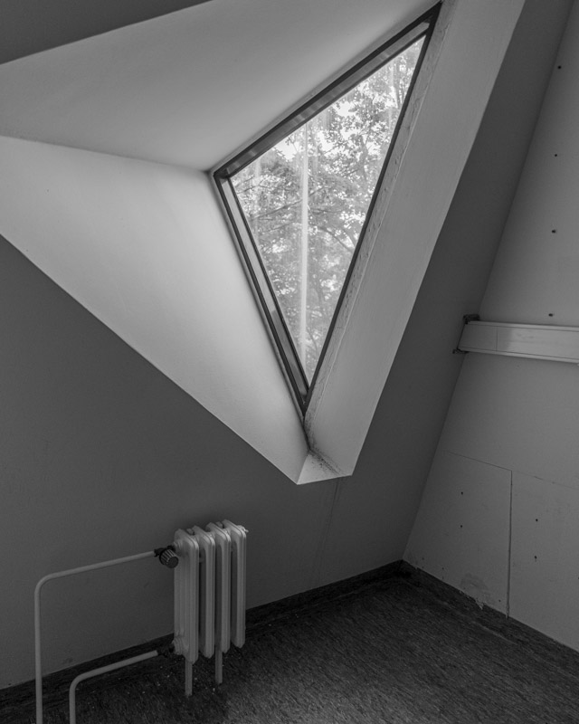 Innenraum im Mäusebunker Berlin: dreieckiges Gaubenfenster mit Blick ins Grüne über einem alten Heizkörper, weiße geneigte Wände – Brutalismus von innen in Schwarzweiß